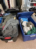 Photo showing two plastic bins filled with assorted worn clothing including vests, shirts, and pants in neutral and muted colors.