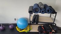 Photo showing six rolled exercise mats on a metal and wood shelf, two purple kettlebells on the floor, and a blue exercise ball on a yellow stand nearby.