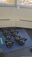 Seven kettlebells arranged on blue padded floor mat in front of a window.
