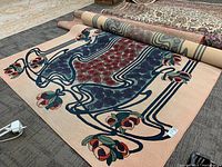 Area rug lying flat on a carpeted floor showing floral and vine pattern with a peach background and partially rolled edges nearby other rugs.