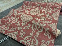 Area rug with red background and cream floral and scroll pattern, partially rolled, lying on a gray carpeted floor indoors with other rugs in background.