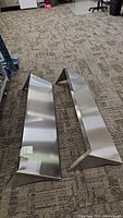 Two angled stainless steel shelf components lying flat on carpeted floor.