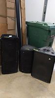 Three black PA speakers of varying sizes in a storage area