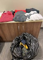 Assorted folded sweaters and sweatshirts laid out on counter with black bag of pants and other clothes on floor.