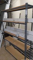 Full view of Uline metal shelving unit with 5 particle board shelves. Shelf is empty except some wiring and cords temporarily draped over shelves.