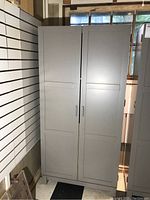 Front view of grey laminate storage cabinet with two doors and black handles standing on feet in a room with slat wall and black and white flooring.