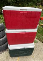 Three large Coleman coolers stacked outdoors, two red and one green, showing front and side views with handles and drainage spouts.