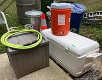 Photo of the lot showing large white Igloo cooler with hinged lid, orange Rubbermaid water cooler with spigot on top, small brown deck storage box with coiled green hose on lid.