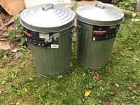 Two Behrens galvanized metal trash bins with lids placed outside on grass, labels present on side showing brand, size, and product features.