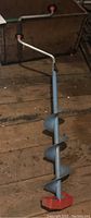 Single coated metal ice fishing auger with blue spiral bit and red base laying on wooden floor, photographed for overall item detail.