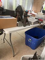 Side view of light colored folding plastic topped table with collapsible metal legs, showing items on top and blue bin underneath.