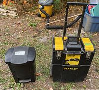 Stanley Mobile Work Center and black waste basket positioned outside on grass and dirt.