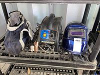 Black and blue Mastercraft welding machine, welding gloves, and blue auto-darkening welding helmet arranged on a black metal shelf.