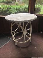 Front angled view of round white painted wicker table with rattan legs and frame, placed on indoor carpet with outdoor garden background.
