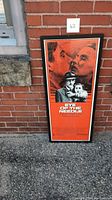 Full view of framed Eye of the Needle movie poster leaning against a brick wall outdoors, showing the entire poster and frame.