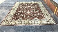 Full view of large brown handwoven Hariz rug with intricate floral and vine patterns and cream borders laid flat on asphalt.