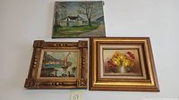 Three oil paintings laid out on a white surface: one landscape of a house, one diver scene in ornate frame, one floral still life in gold frame.