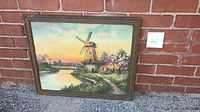 Framed art print showing full scene of windmill, cottage, waterway, and sunset sky leaning against a brick wall.
