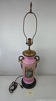 Full view of the vintage porcelain lamp, showing the pink body, black circular base, electrical cord, and top brass finial and fittings.