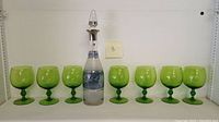 Full view of a clear crystal decanter with silver bands and stopper, surrounded by eight green stemmed wine glasses.