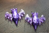 Pair of purple translucent plastic bat earrings lying on a black leather surface, showing front details including wings and body spots.