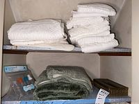 White and beige bath towels folded and stacked on a shelf with folded green/gray bath rugs below, as well as travel bottles set and decorative tissue box cover on bottom shelf.