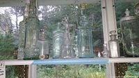 Front view of nine assorted decorative glass bottles and decanters on a shelf in front of a window. Various shapes and colors including clear and light green glass, cork and glass stoppers.