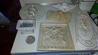 All 7 items together: 6 cherub-themed wall plaques and one small square cherub pot on a table showing their size and style.
