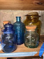 Set of four colored glass jars and one apothecary bottle on a wooden shelf.
