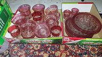Two cardboard boxes holding the entire lot of rose colored glassware displayed on a patterned rug. One box contains stacked plates and goblets, the other contains dessert cups and smaller glasses.