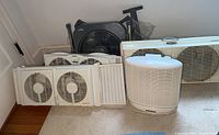 Photo showing seven assorted fans and air circulators in a line on floor against wall.