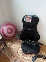 Photo showing a black Pro Shiatsu massage seat with a user manual on top and large pink and smaller green and blue exercise balls placed beside it on a patterned rug and wooden floor.