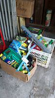 Photo showing cardboard and plastic crate boxes filled with assorted garden care supplies including pest control sprays, nutrients, gardening tools, and plant markers.