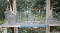 Full shelf view showing assorted glassware including candy jars, stemware, canisters, and vases against window background
