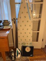 Complete view of Seymour ironing board with floral cover, Health O Meter scale, and both irons placed on floor in front of a door.