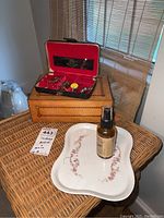 Photo of two jewelry boxes stacked, white porcelain tray and amber spray bottle on woven surface.