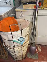 Two canvas laundry baskets stacked in metal frame with orange mesh hamper inside, next to Euro-Pro steam mop positioned upright on red floor in laundry area next to washing machines.