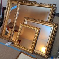 Four gold-toned framed mirrors arranged on the floor, showing front reflective surfaces and frame details with varied frame styles and sizes.