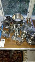 Overview of the pewter tableware set on a wooden table, showing bowls, mugs, pitcher, candlesticks, butter dish, plates, and other items.