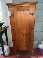 Full front angled view of pine wood pantry cabinet showing door and wooden peg latch, cabinet standing on red floor beside small table.