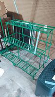 Front angled view of the green metal folding plant stand showing three slatted shelves and structural frame, set indoors on concrete floor.