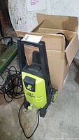 Sun Joe power washer unit positioned next to a cardboard box on a concrete floor, showing front and side details including handle, wheels and hoses.