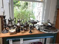 Overview of silverplated serving ware set displayed on wooden table by window, includes pots, bowls, platters, candlesticks.