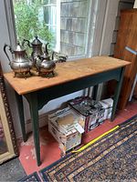 Full view of pine wood console table showing top with stain marks and decorative silver teapots on top. Green legs and side panels visible. Located against windowed wall on porch area.