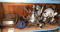 Photo of tea ware set on wooden shelf including silver tone teapot, ceramic floral teapot, blue cast iron teapot, sugar and creamer set, and small covered container.