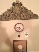 Full view of the carved lion head plaque, wall clock with pendulum beneath it, and framed relief art below the clock.