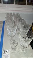 Multiple clear Waterford Crystal stemmed wine glasses with intricate cut patterns, placed on a white table, along with a small Waterford Crystal vase.