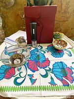 Overview of lot items on floral printed tablecloth including the coin, bread basket, document portfolio, wine opener, and bowls
