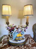 Full lit view of the porcelain figurine lamp on a brass base with floral porcelain roses as decoration under the lamp shades.