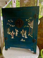 Full front view of the blue lacquered cabinet showing the detailed semi-precious stone inlays and relief work of Asian figures and floral motifs.